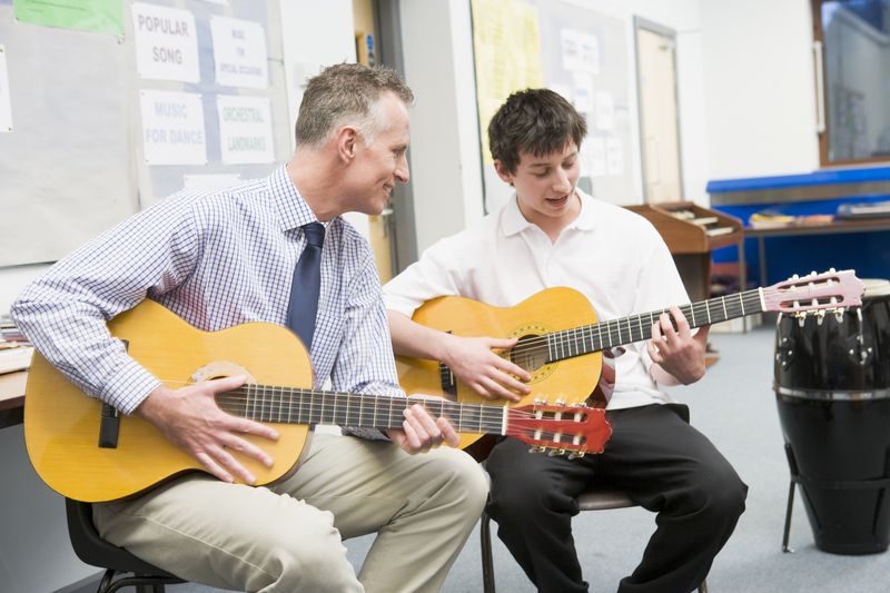 Junge spielt Gitarre bei Gitarrenunterricht neben Gitarrenlehrer im Klassenraum